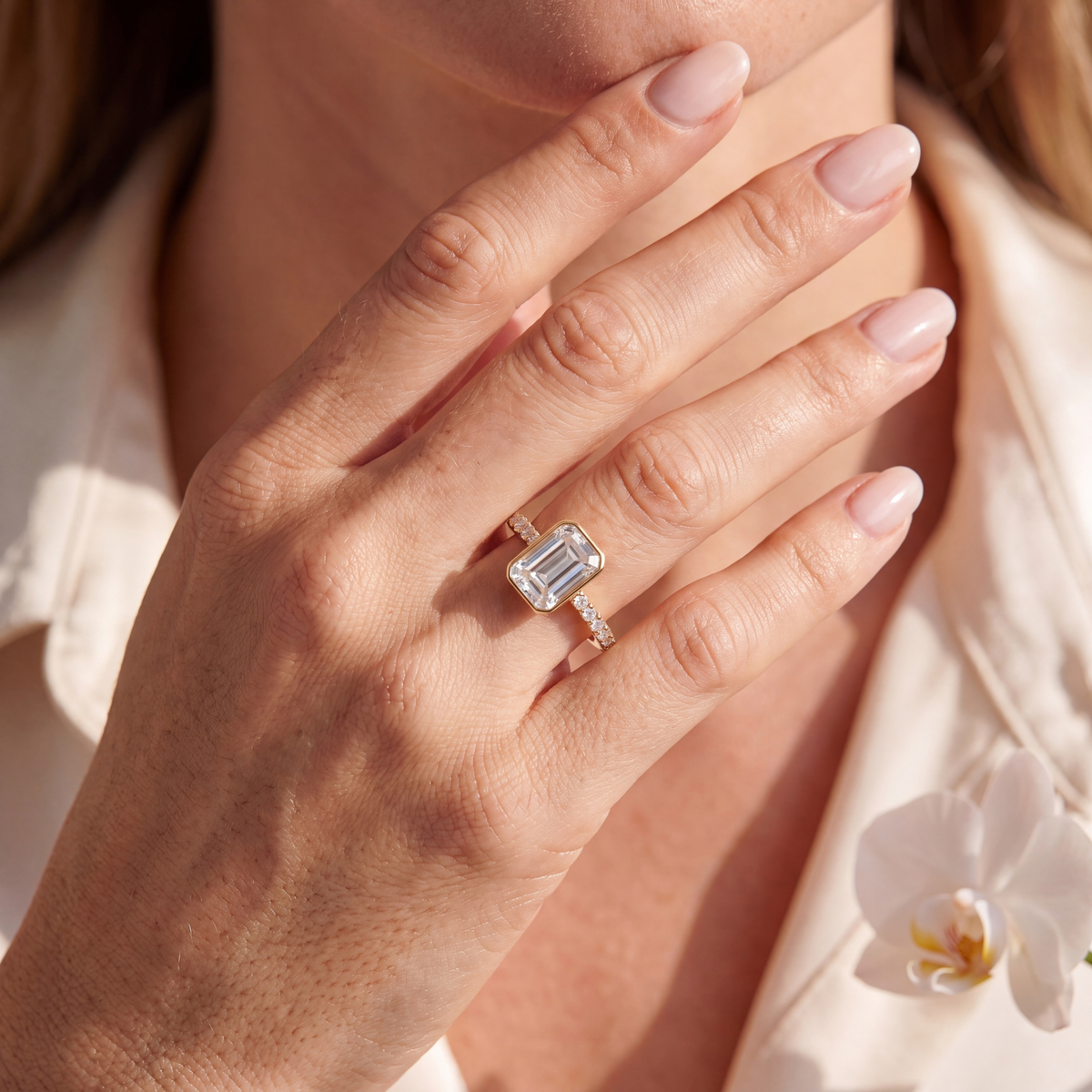 Emerald-cut bezel moissanite engagement ring with a pavé band in yellow gold worn on a hand.