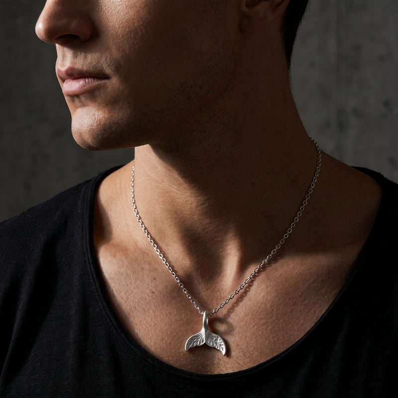 Man looking left while wearing a silver whale tail pendant necklace over a black shirt.