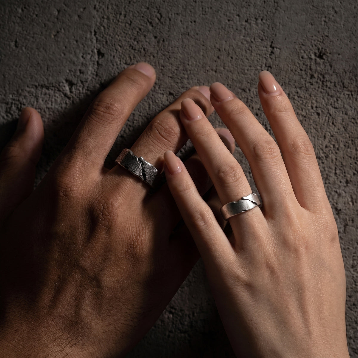 Two hands wearing silver open rings with textured fissure details over a gray textured surface.