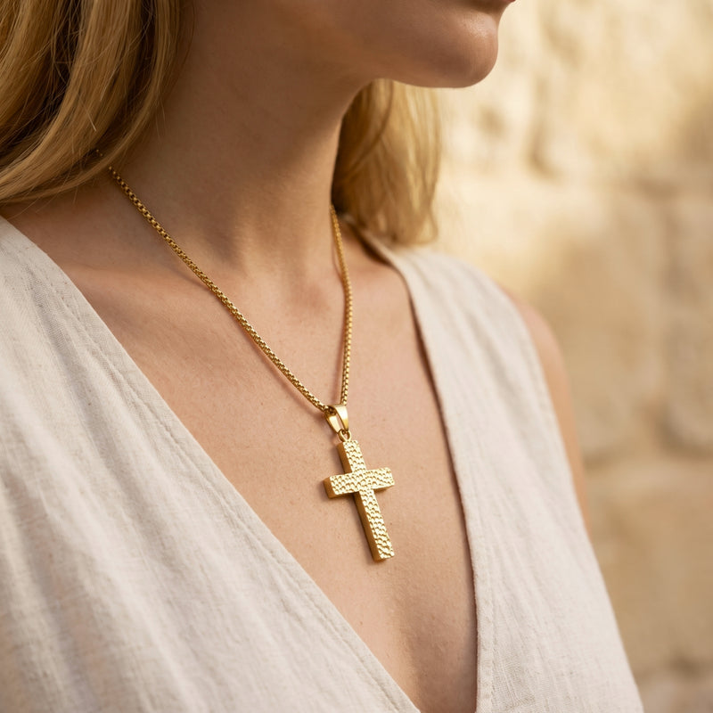 Woman wearing a gold-tone hammered cross pendant necklace over a light-colored open top.