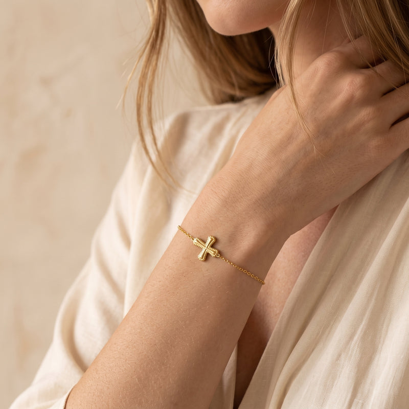 Model wearing a gold-toned cross bracelet on her wrist with her hand near her neck.