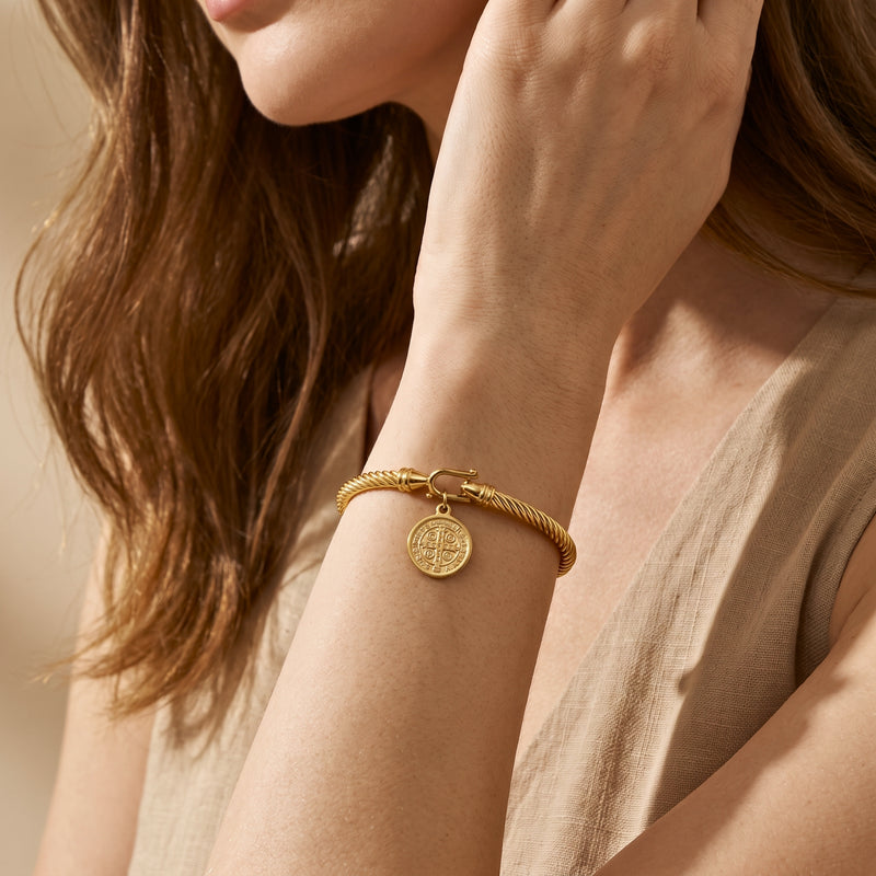 Woman wearing a gold-tone twisted cable bracelet with a dangling medal charm on her wrist, dressed in beige.
