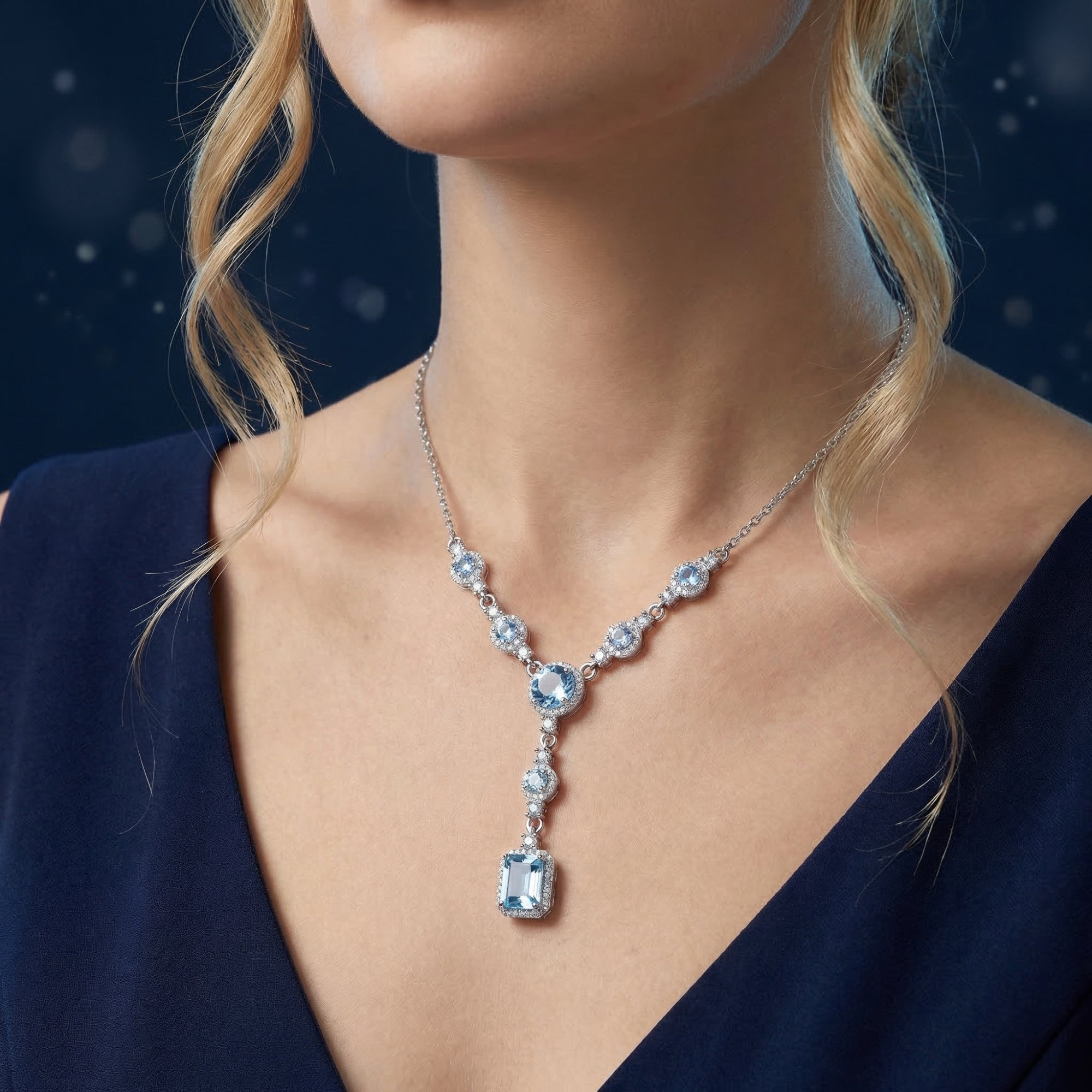 Lorelei Cascade necklace with emerald cut blue topaz and white gold worn by a model in a dark blue dress.