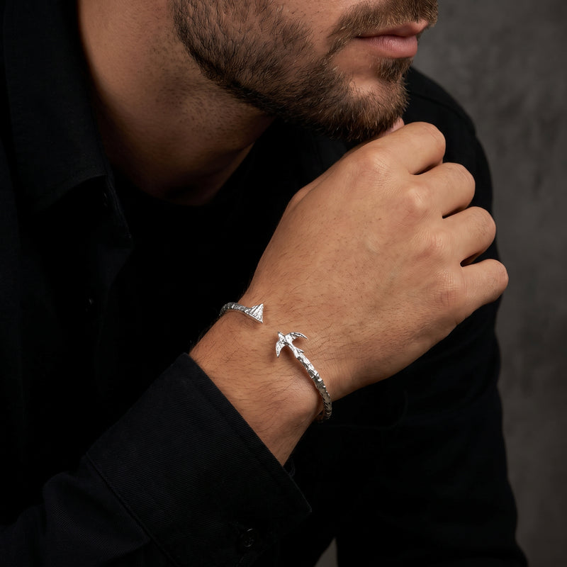 Silver open cuff bracelet worn on a man's wrist, viewed near his chin with mountain and bird ends visible.