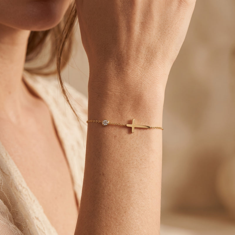 Close-up of the gold-tone sideways cross bracelet with a round clear stone worn on a woman's wrist.