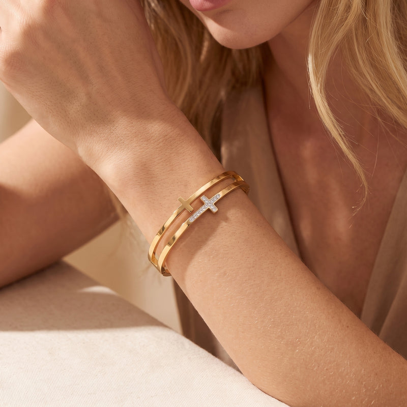 Model wearing the gold-tone double-band cross bangle on a raised wrist beside her chin.