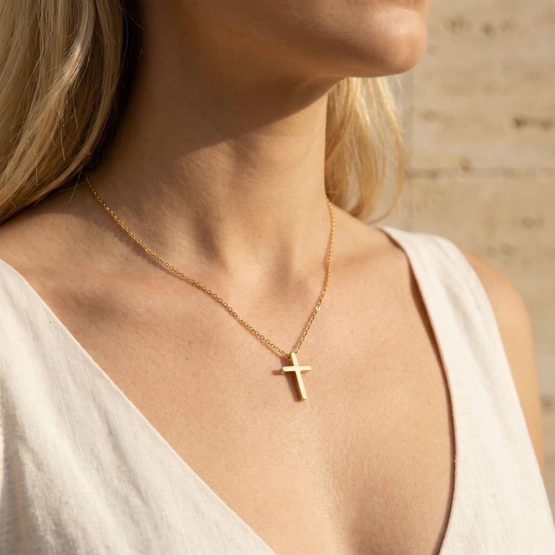 Close-up of a woman wearing a gold-tone cross necklace with a thin chain over a white sleeveless top.