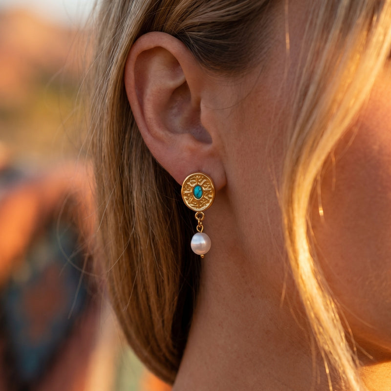 Gold sunburst drop earring with turquoise cabochon and pearl worn on a woman's ear in warm outdoor light.
