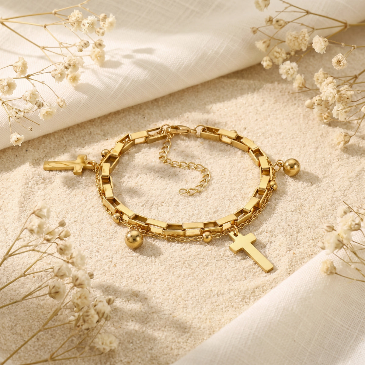 Gold double-layer cross bracelet laid open on sand with the clasp and extender chain visible.