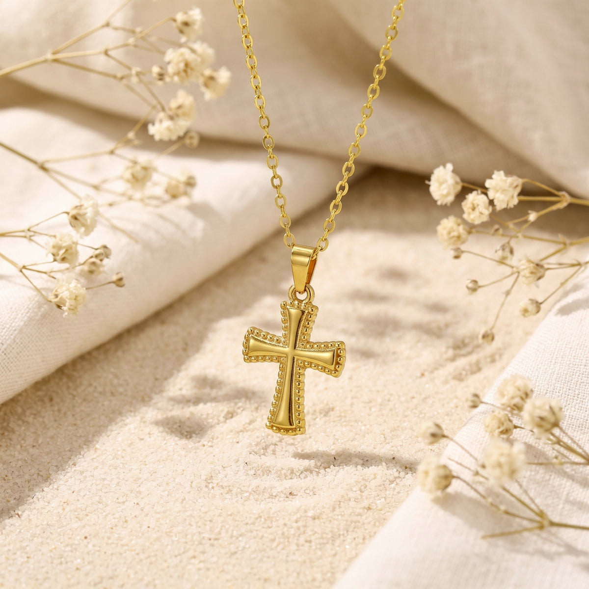 Gold-tone beaded cross pendant hanging vertically above sand with surrounding dried floral accents.
