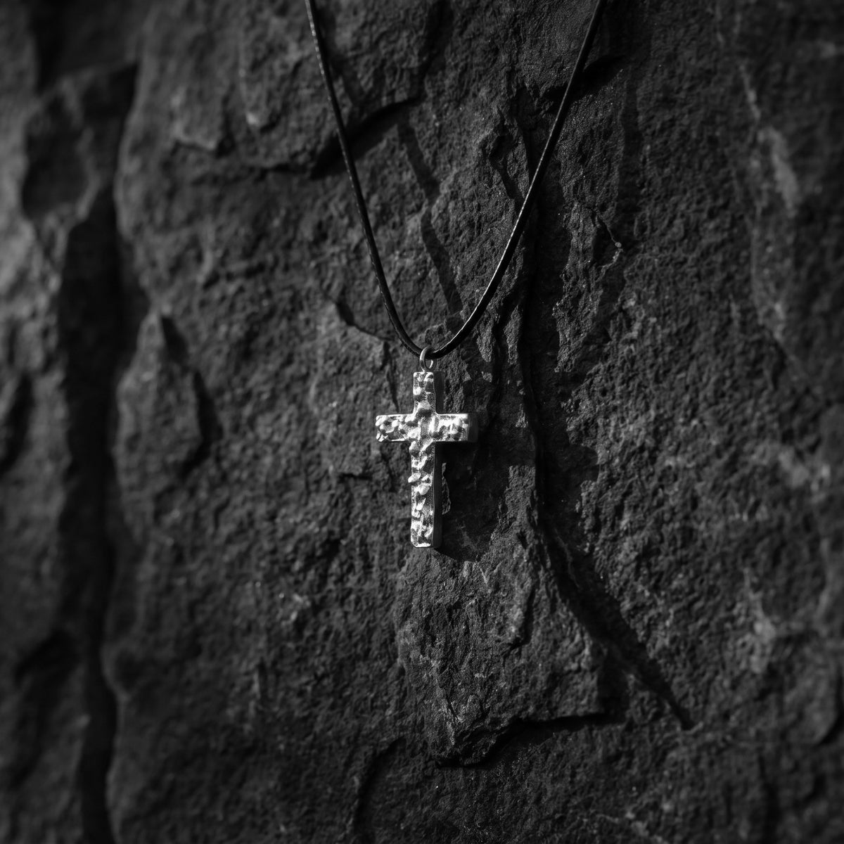 Textured silver cross pendant hanging vertically on a black cord against a rough dark rock surface.