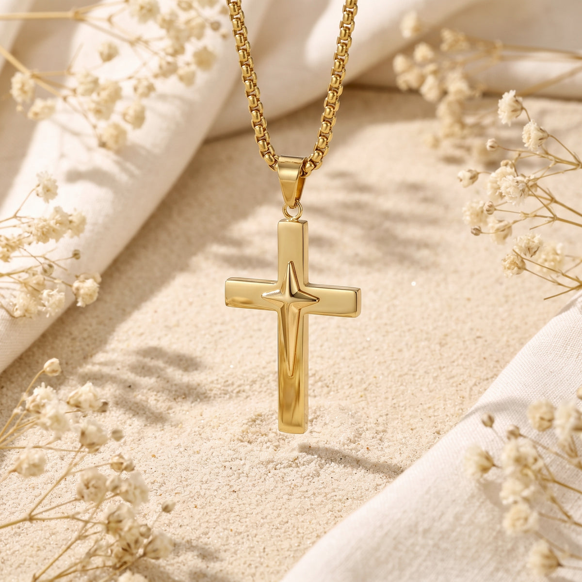 Close-up gold-tone star cross pendant hanging over sand, framed by white fabric and dried flowers.
