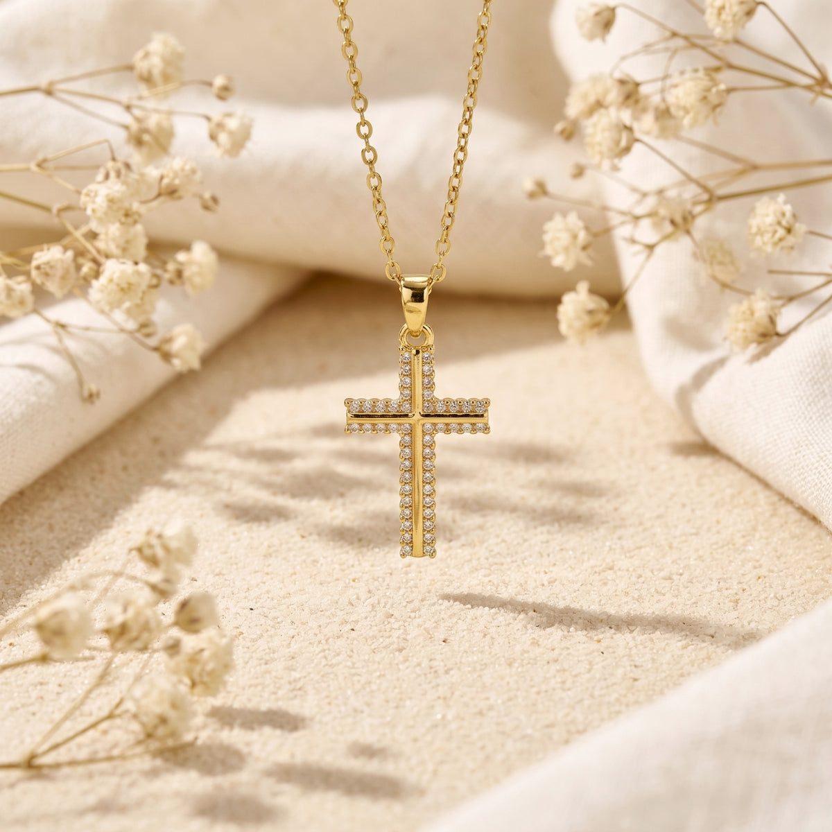 Gold-tone pavé cross necklace hanging vertically over sand with dried flowers.