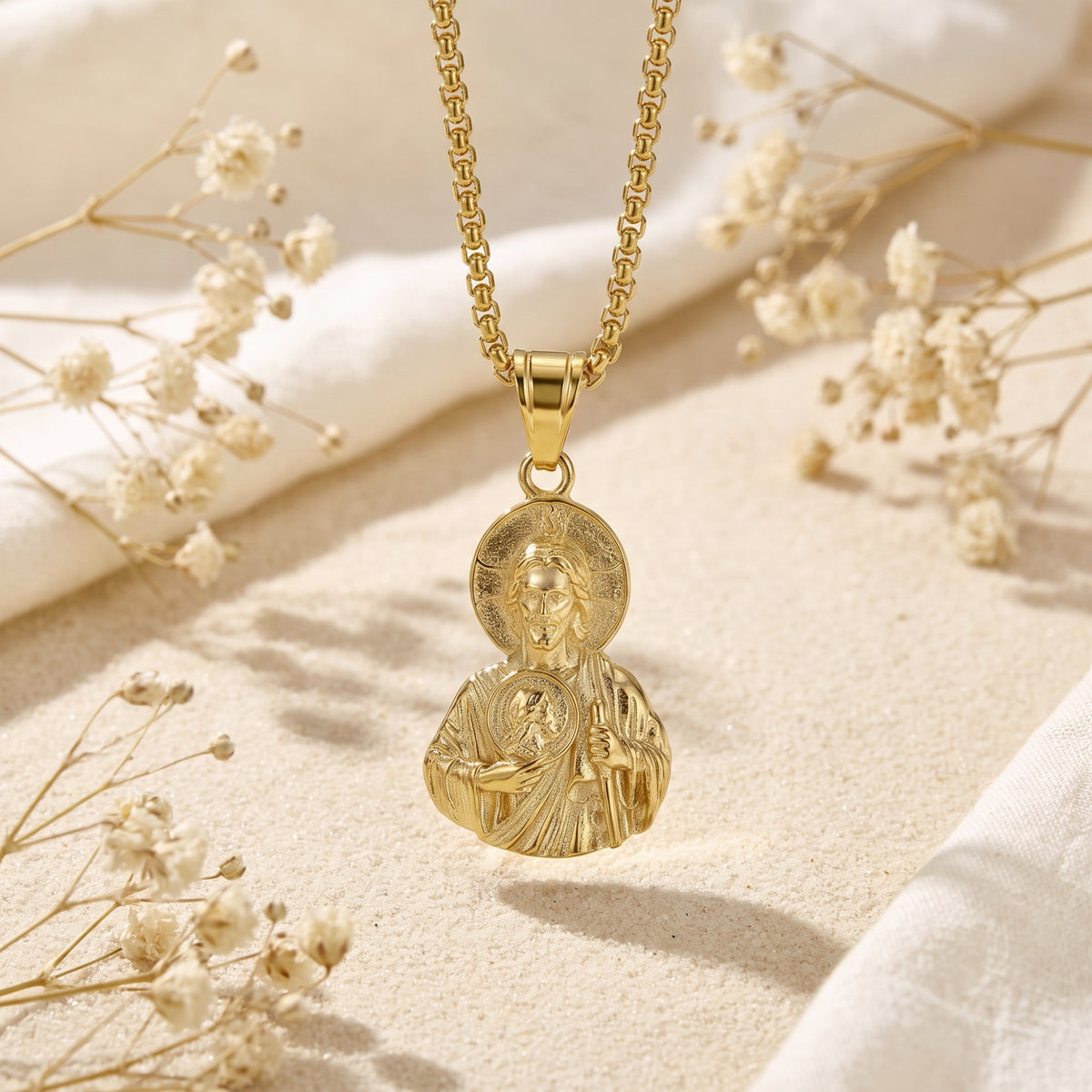 Front view of the gold finish sacred icon pendant hanging from a box chain above light sand.
