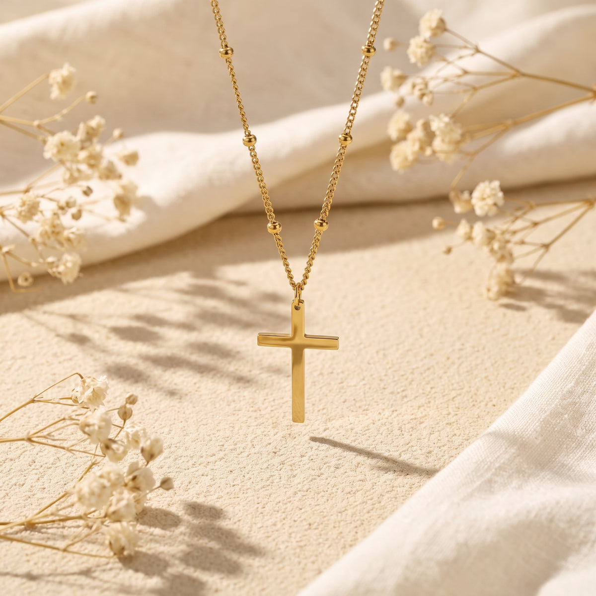 Gold-tone cross necklace suspended above sand with dried white flowers surrounding it.