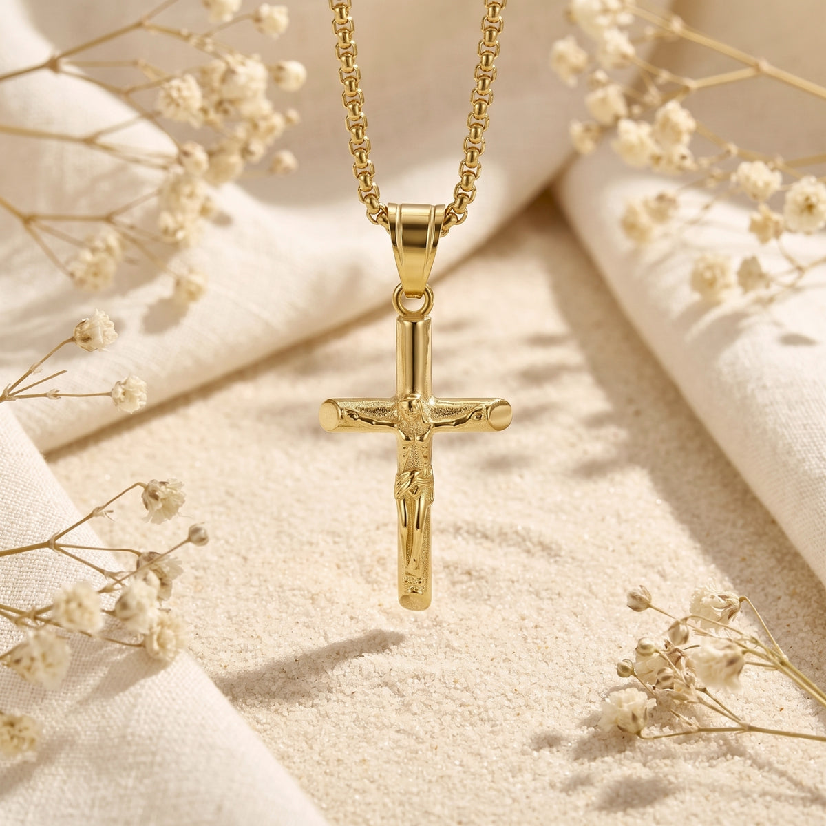 Gold-tone crucifix pendant hanging vertically above sand, with white fabric and dried flowers around it.