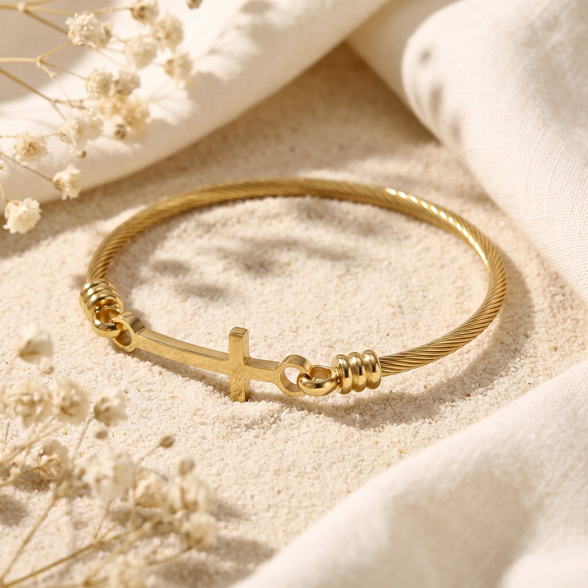 Angled flat-lay view of the gold-tone twisted cable bracelet with a cross on pale sand and cream fabric.
