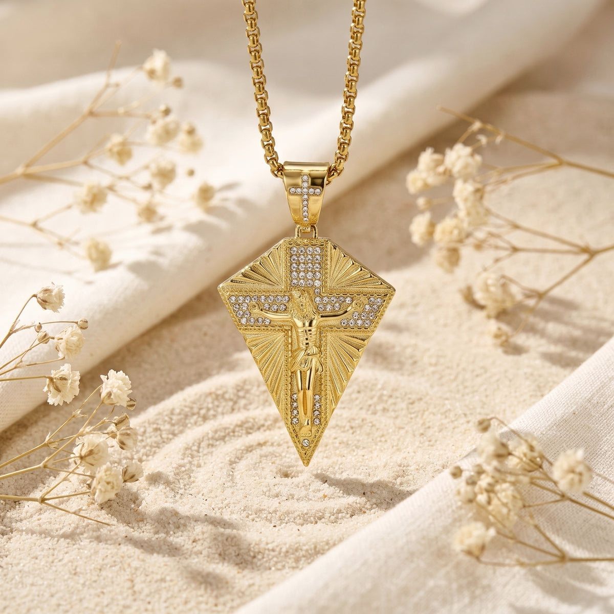Gold-finished crucifix pendant necklace suspended above sand, with dried white flowers in the background.