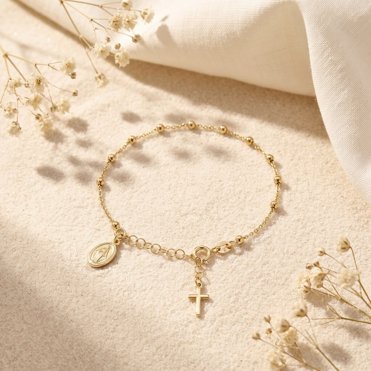 Overhead view of a gold beaded charm bracelet with medal and cross on sand among dried flowers.