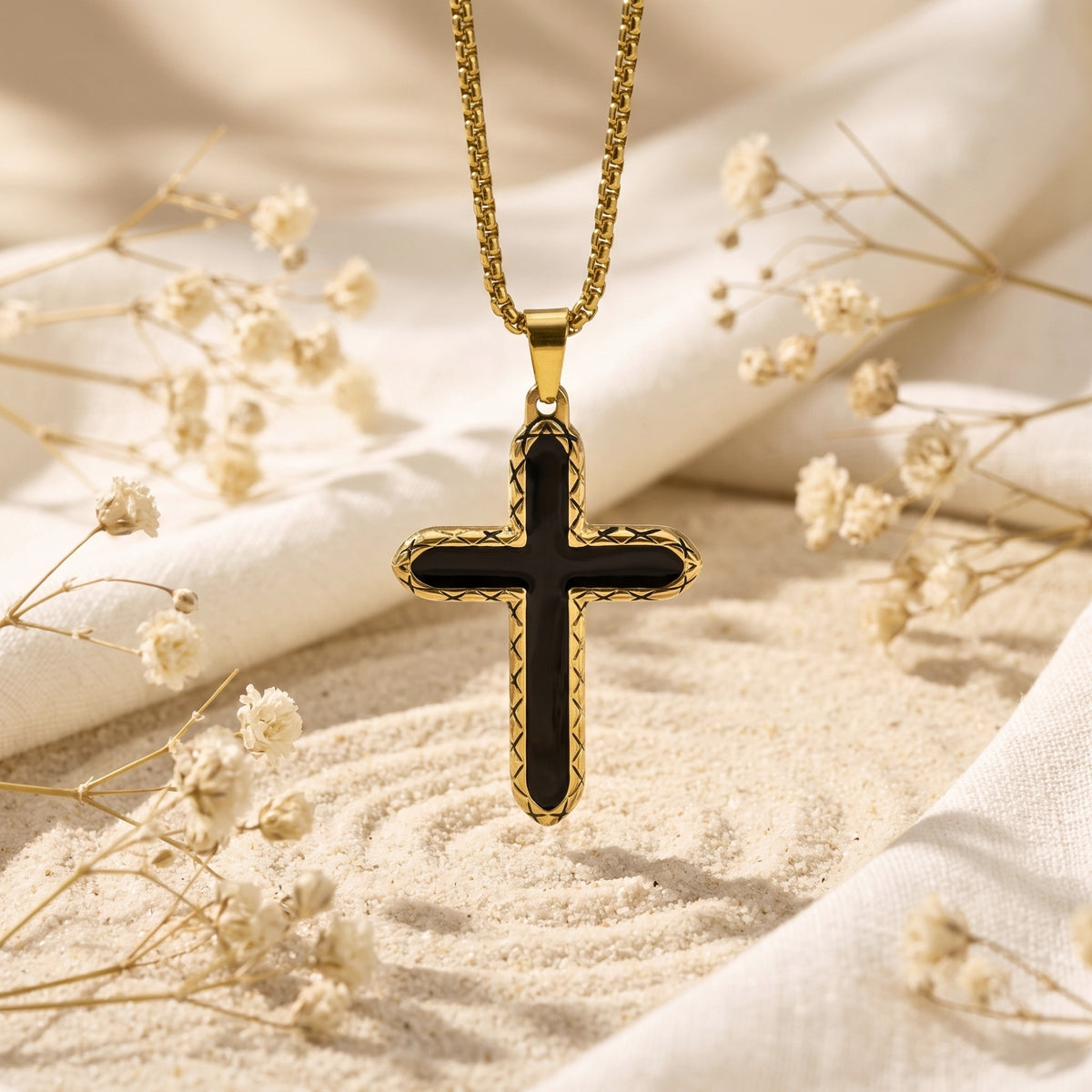 Gold-tone black-inlay cross necklace hanging vertically above sand, framed by white fabric and dried flowers.