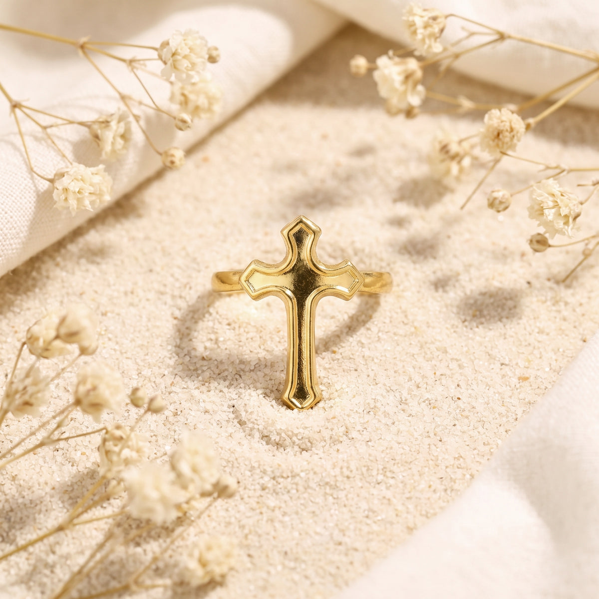 Front view of a gold-tone cross ring standing upright in sand among cream fabric and dried flowers.