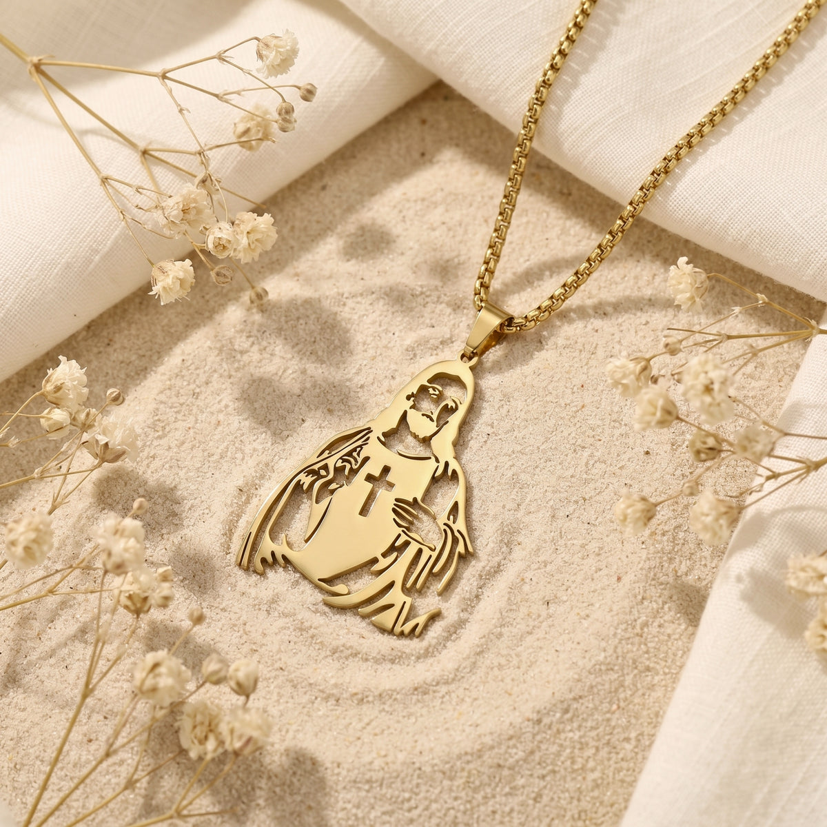 Gold-tone Christ silhouette pendant with a cross cutout lying on sand among white linen and dried flowers.