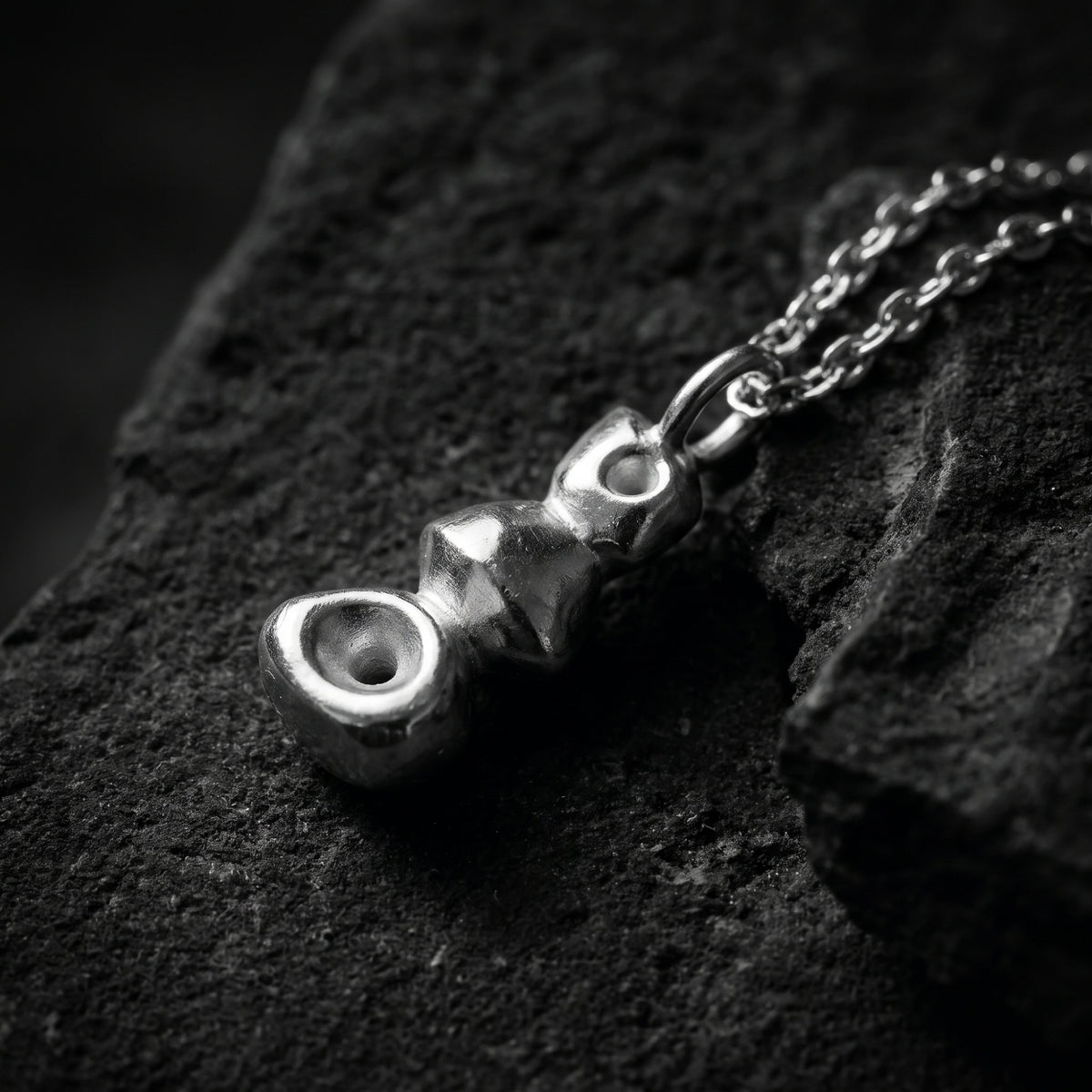 Macro close-up of the silver stacked cairn pendant resting diagonally on dark rock.