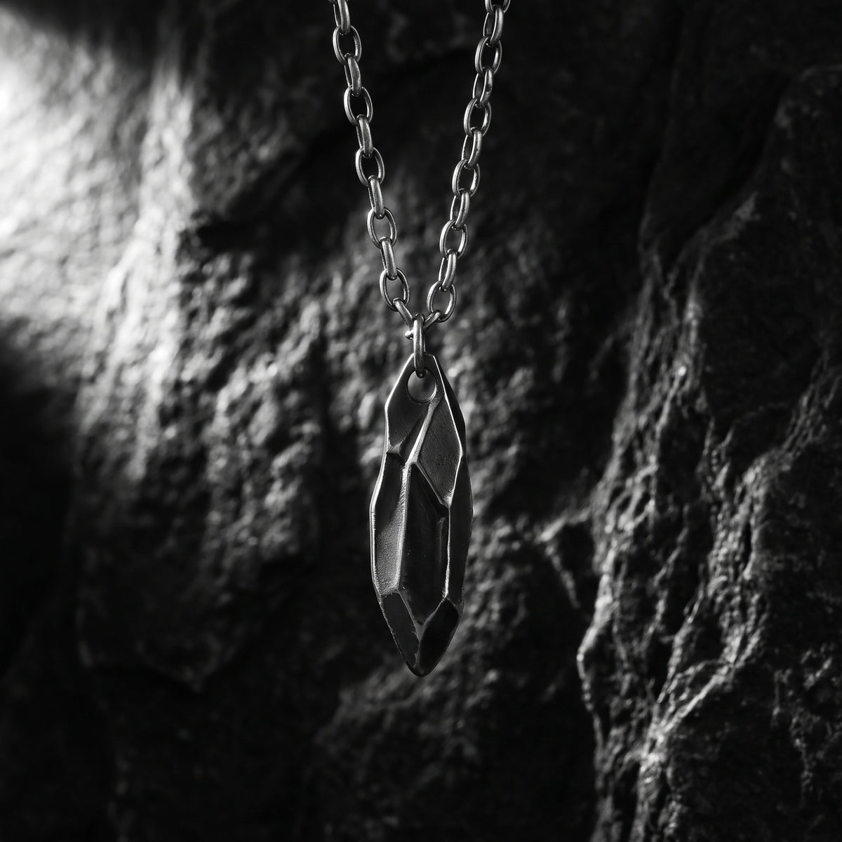 Faceted silver shard pendant hanging vertically against a dark rock background.
