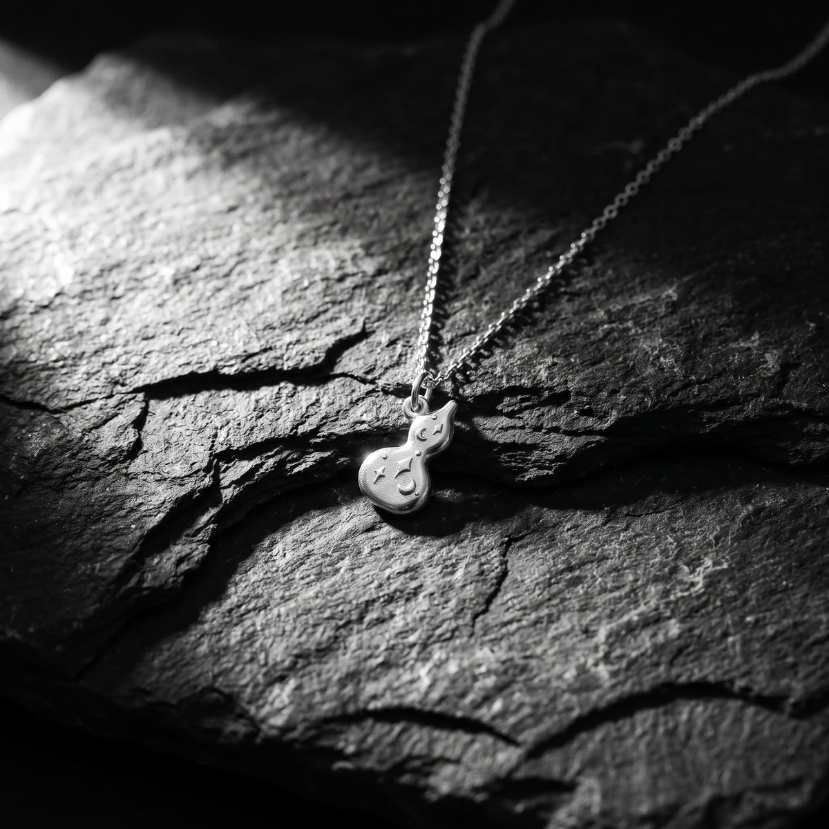 Silver gourd pendant lying flat on textured dark slate, with its chain extending upward.