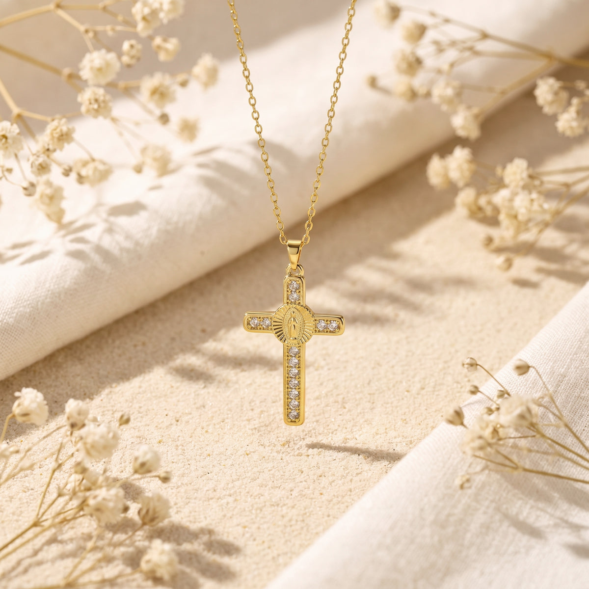 Gold-tone cross necklace laid flat on sand and fabric, with a central Virgin Mary medallion and clear accent stones.
