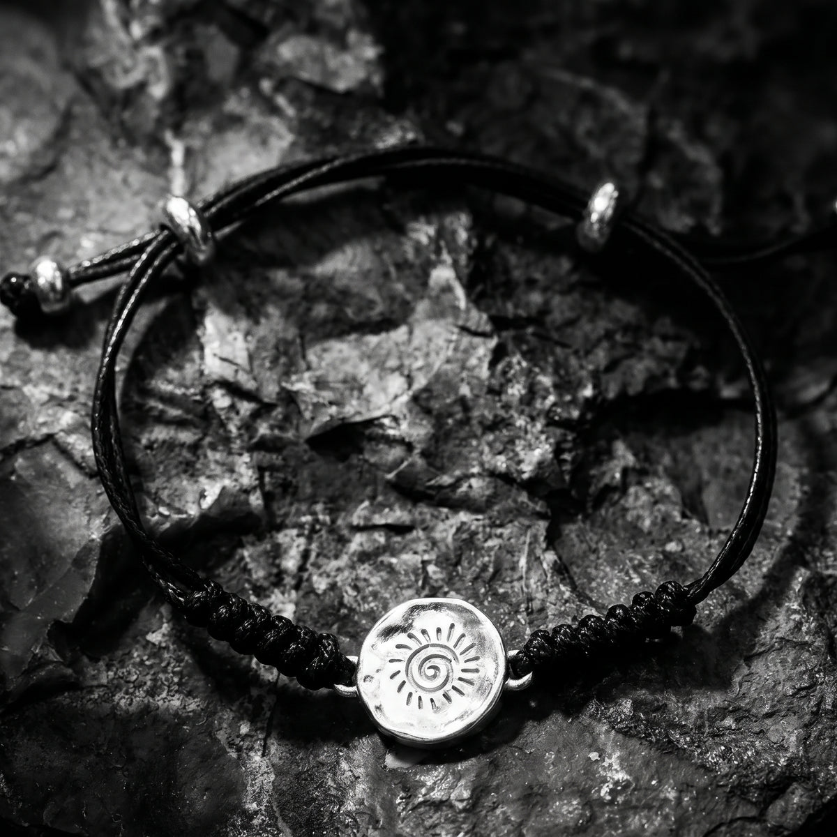 Full black cord bracelet with a round silver sun medallion and sliding knots on jagged rock.