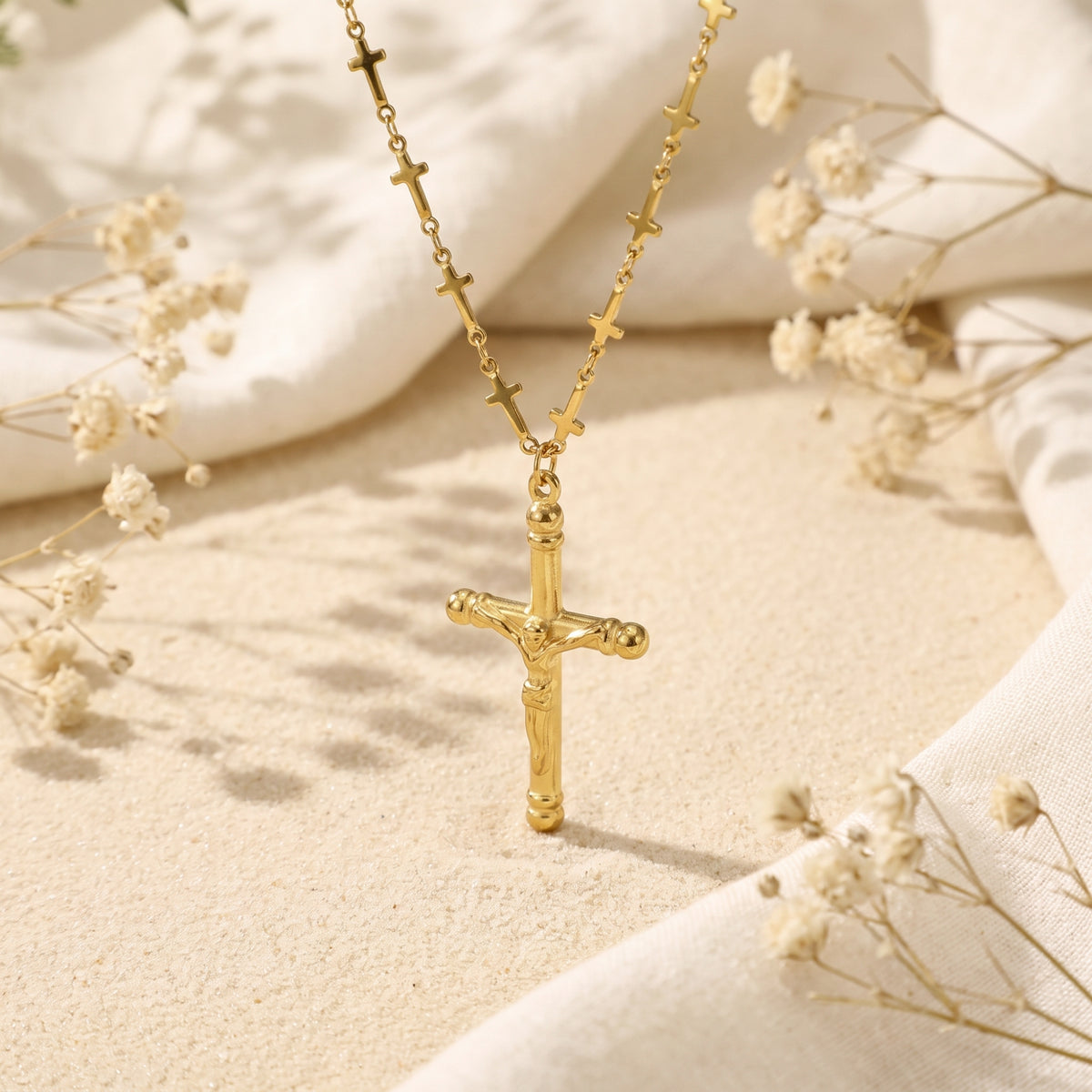 Gold-tone crucifix pendant hanging upright over sand, with small cross stations visible along the chain.
