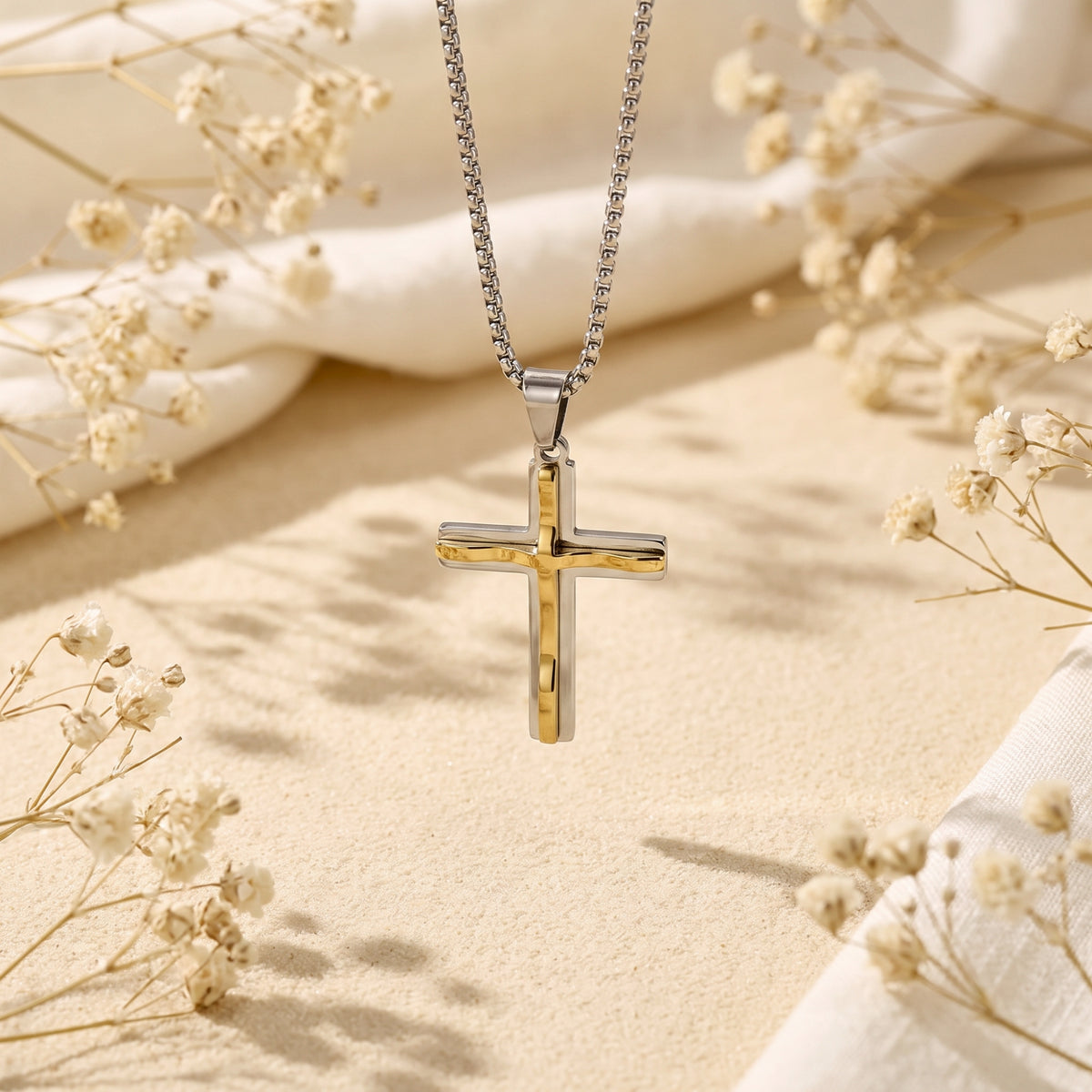 Two-tone silver and gold cross pendant suspended vertically above sand with dried floral accents.