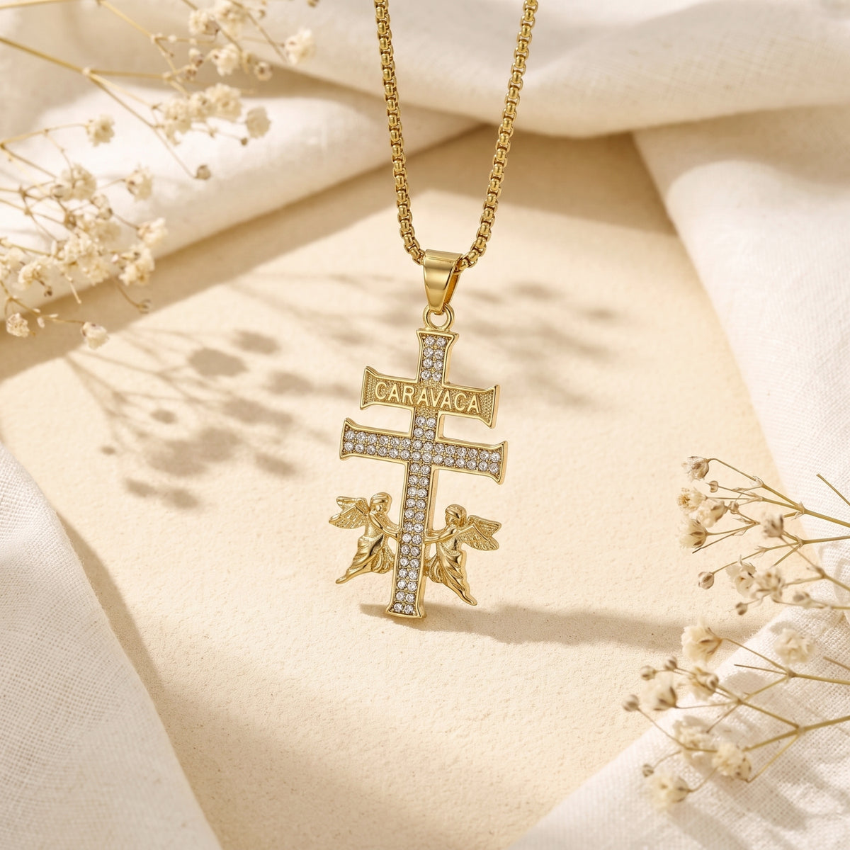Gold Caravaca double-bar cross pendant with sparkling stones suspended over cream fabric.