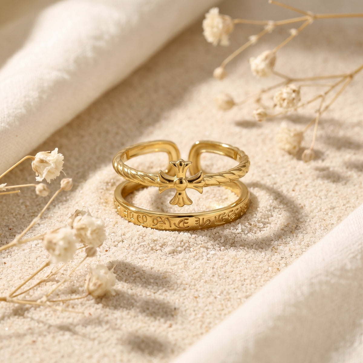 Front view of a gold-tone double-band cross ring arranged on sand, showing the cross and engraved script.