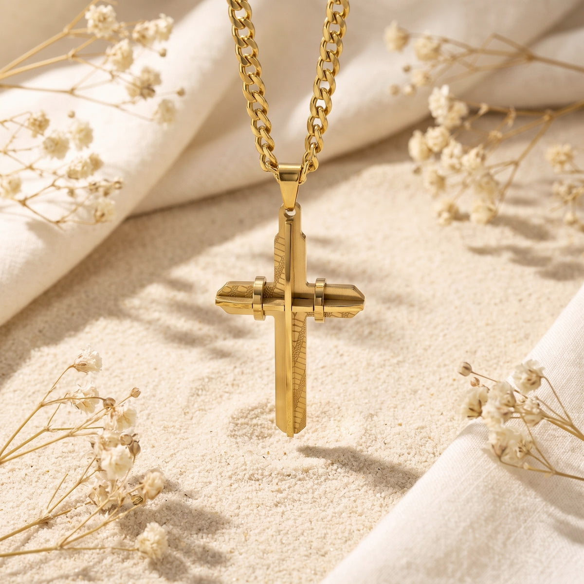 Front view of a gold-toned layered cross pendant hanging over pale sand and dried white flowers.