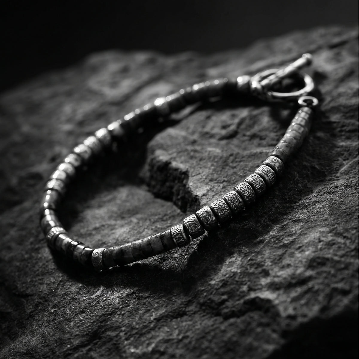 Angled close-up of the silver bracelet on dark rock, focusing on carved cylinder links with the clasp in the background.