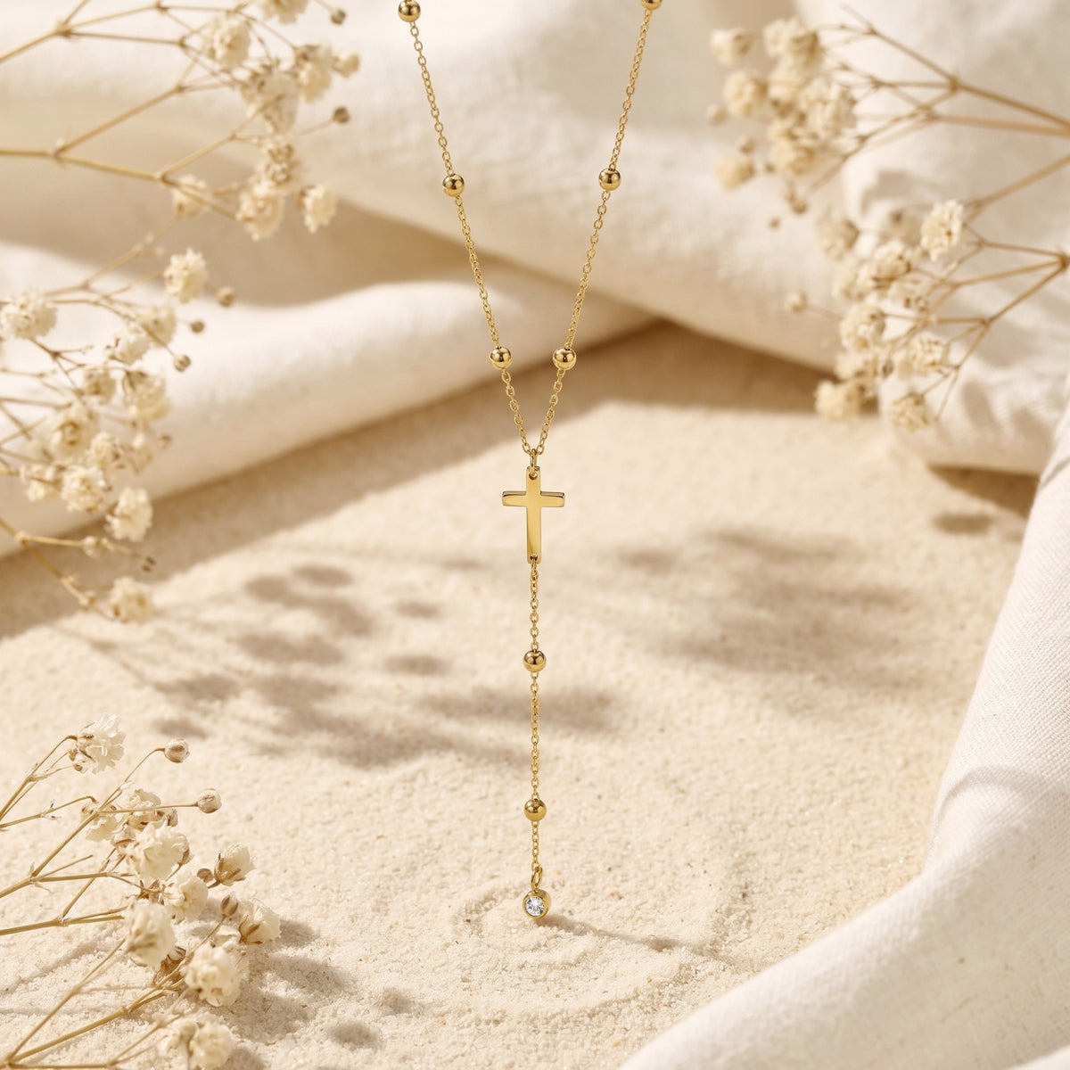 Gold-tone cross lariat necklace displayed vertically over sand among dried white flowers, with bead stations