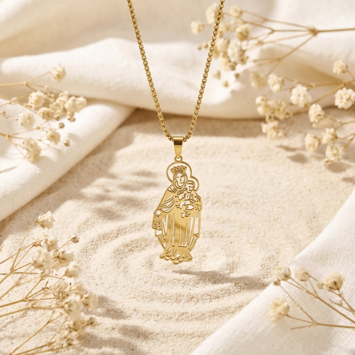 Gold Madonna and Child pendant necklace hanging vertically over sand, white fabric, and dried flowers.