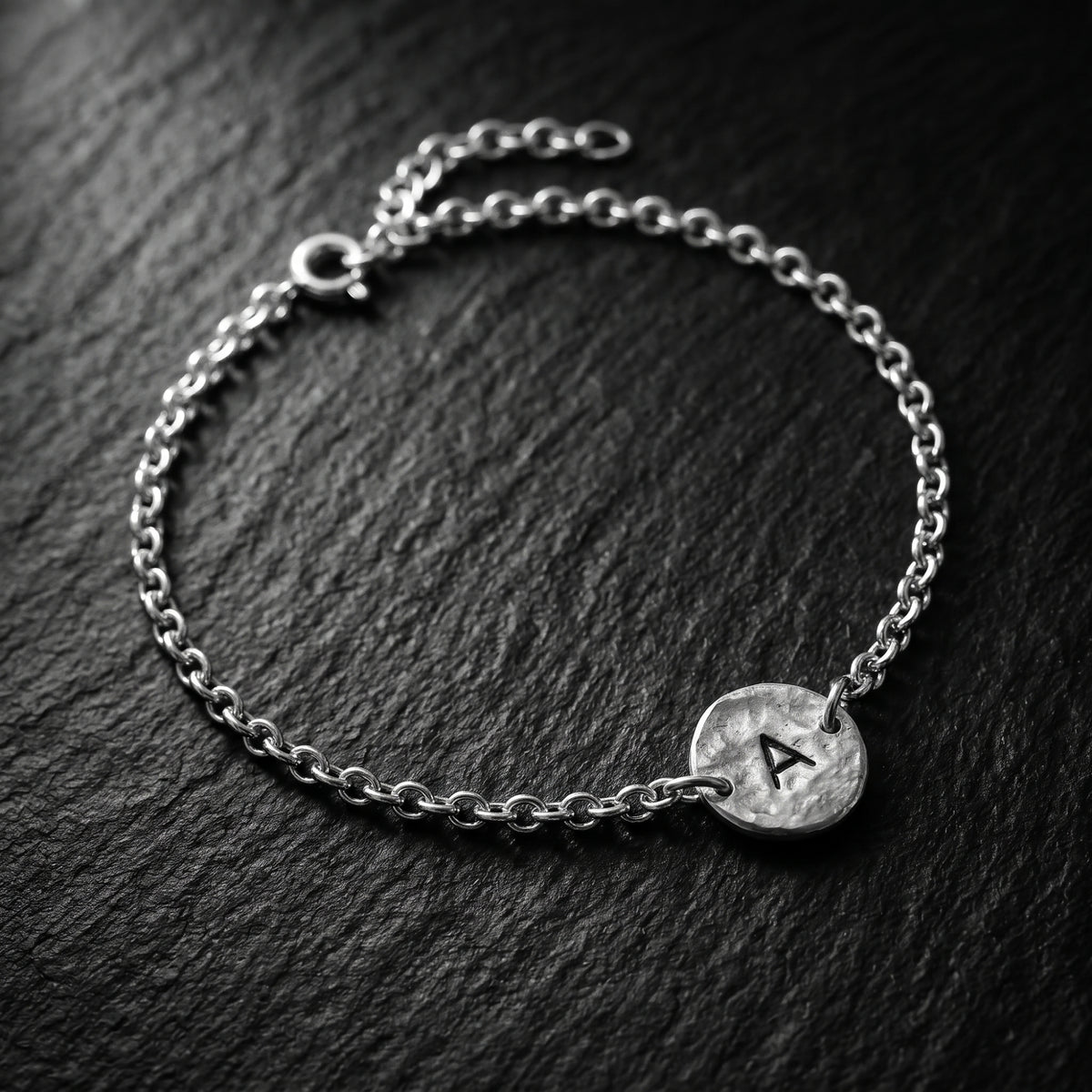 Angled view of a silver initial A bracelet on a textured dark background, showing the disc, clasp, and extension chain.