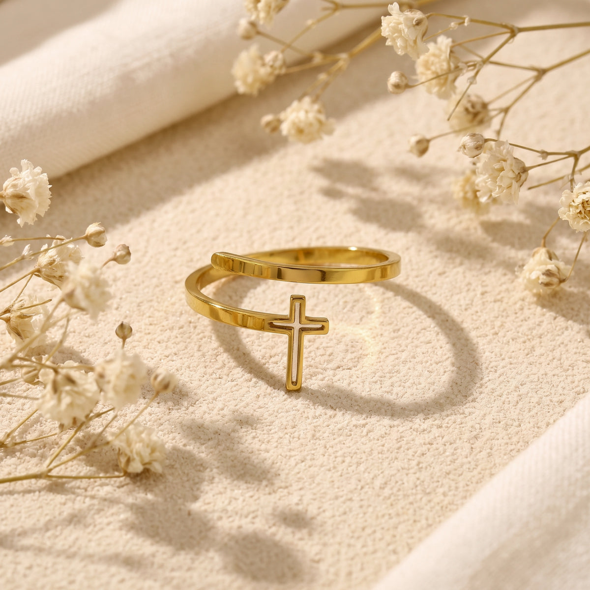 Gold-finish open-wrap cross ring resting on sand among dried white flowers.