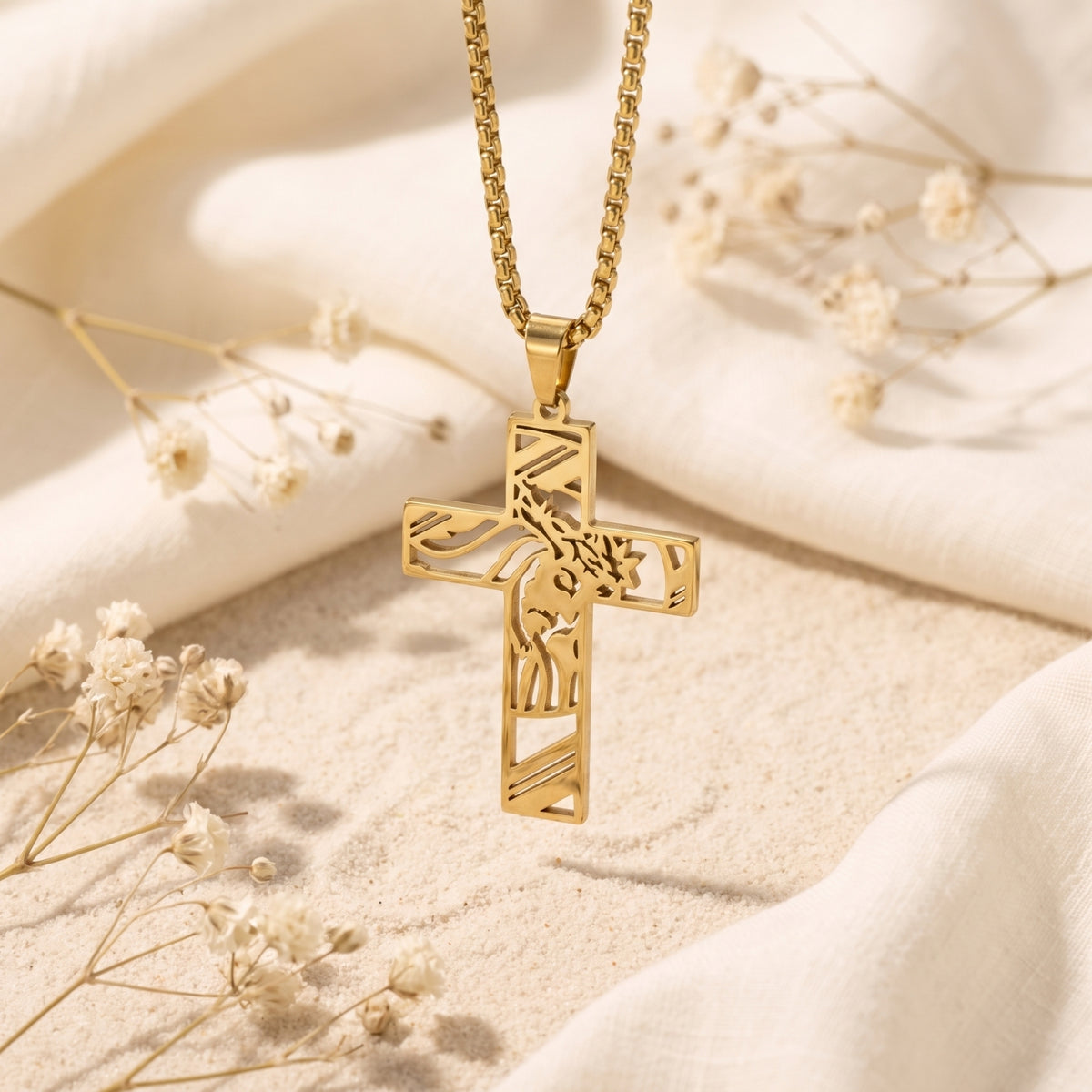 Gold-tone sculpted cross pendant hanging vertically over sand, white fabric, and dried flowers.