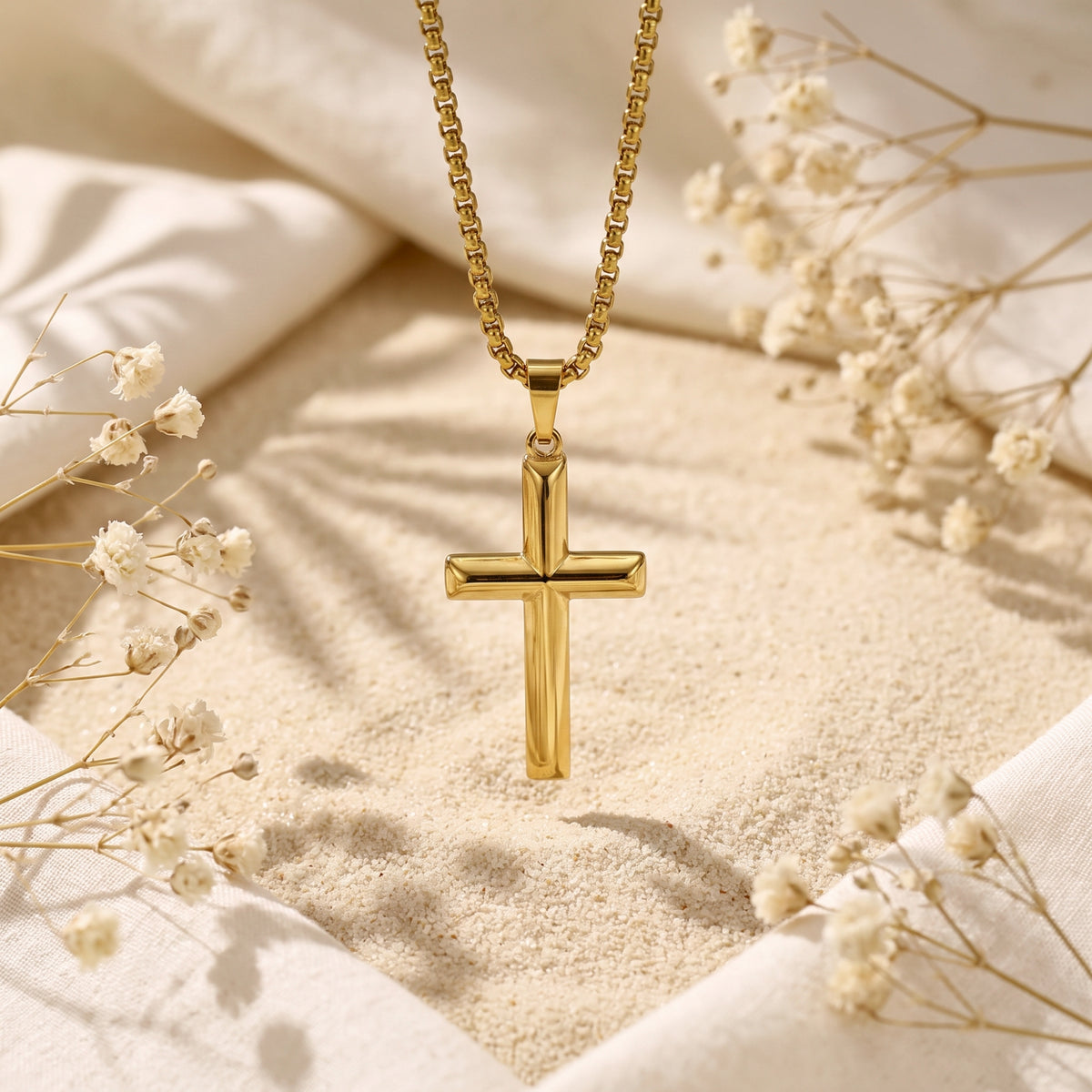 Gold-tone cross pendant hanging vertically above light sand with dried floral accents.
