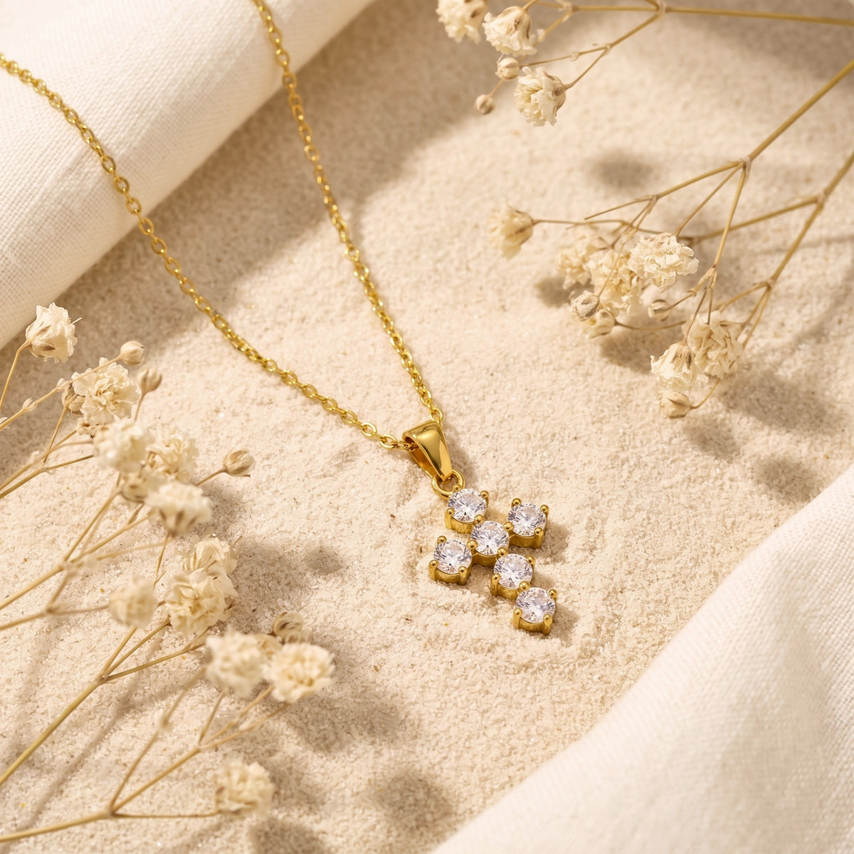 Angled flat lay of the gold-tone cluster cross necklace on textured sand beside dried flowers
