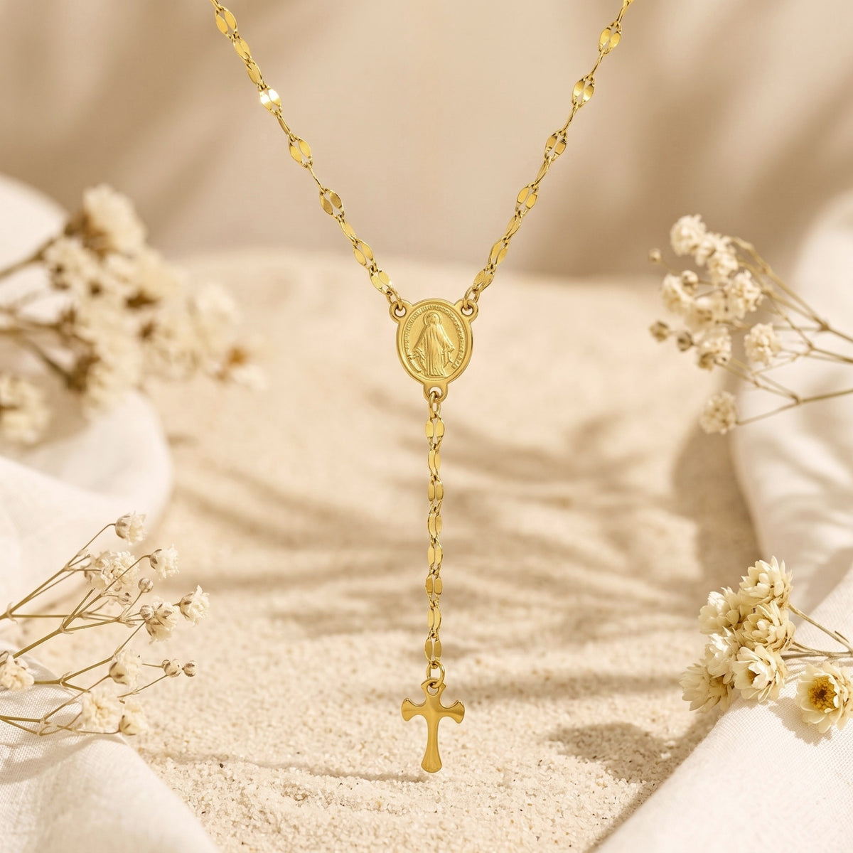 Centered view of the gold-tone rosary necklace hanging vertically over a sandy backdrop with dried flowers.