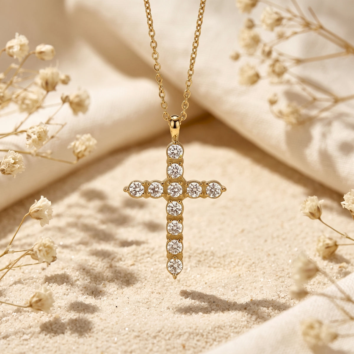Front view of a gold-tone cross pendant with round clear cubic zirconia stones hanging over sand.