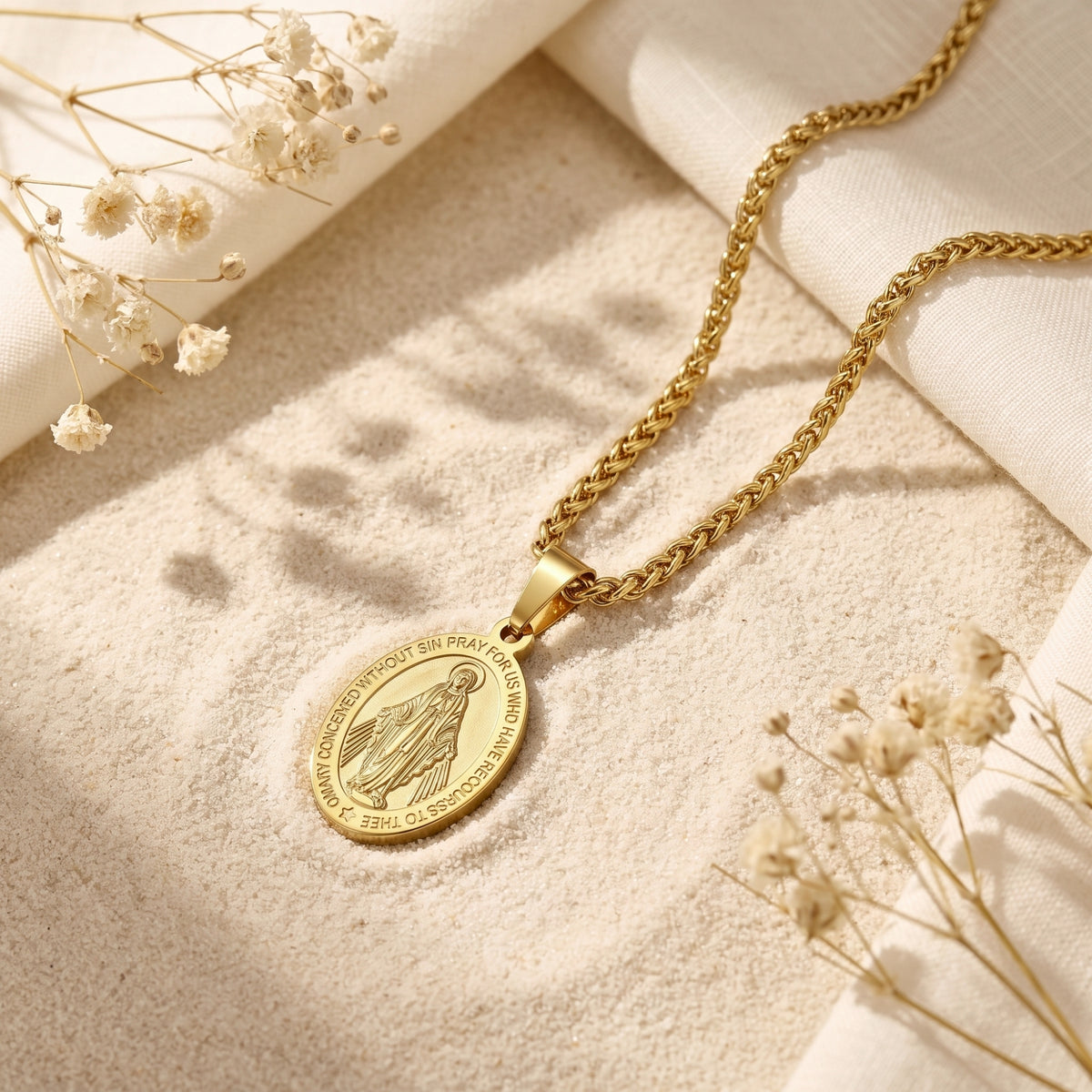 Gold-tone Miraculous Medal necklace laid diagonally on sand among dried white flowers.