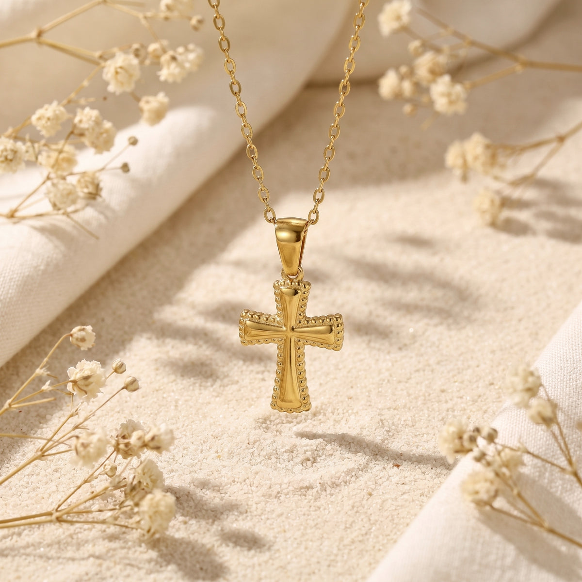 Suspended gold-tone cross pendant necklace with a beaded border against sand and dried flowers.