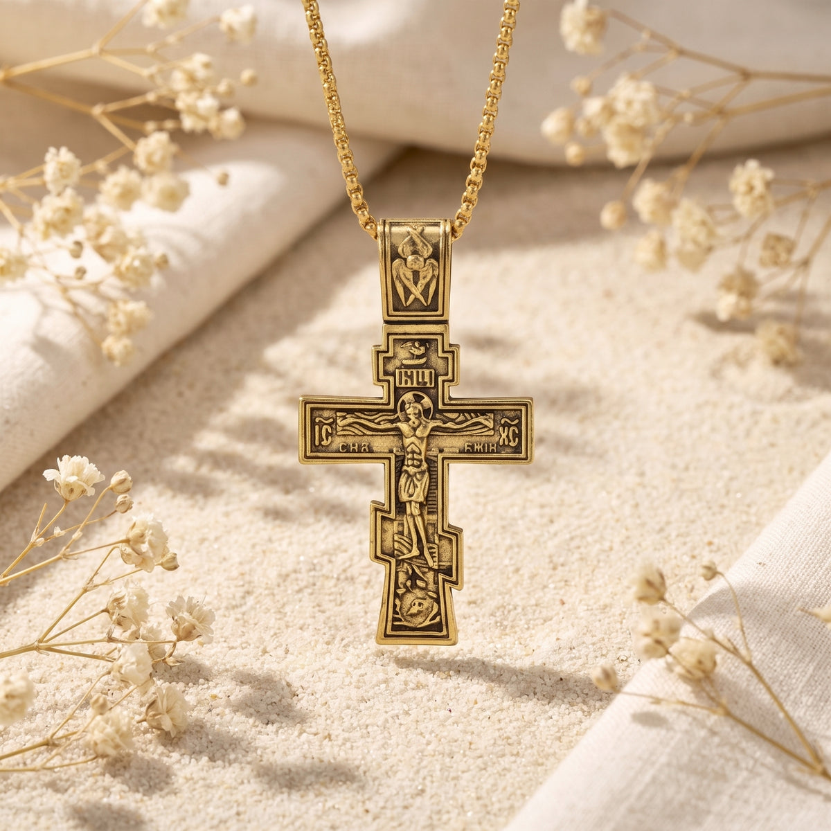 Front view of a gold-tone Eastern cross pendant hanging above pale sand, with white dried flowers in the background.