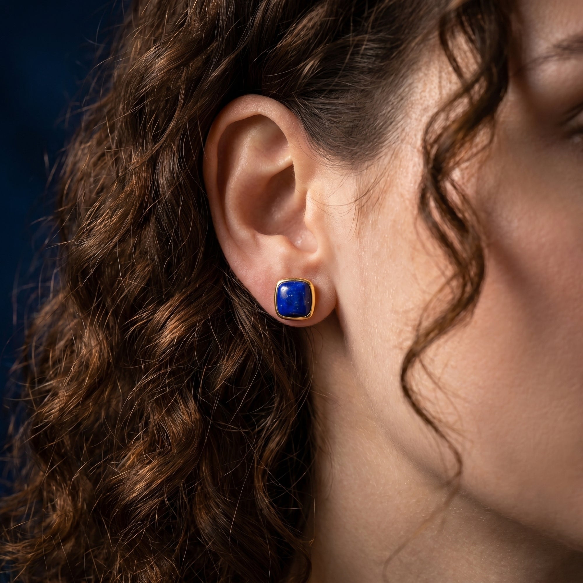 Woman with curly hair wearing a cushion cut natural lapis lazuli stud earring in yellow gold.